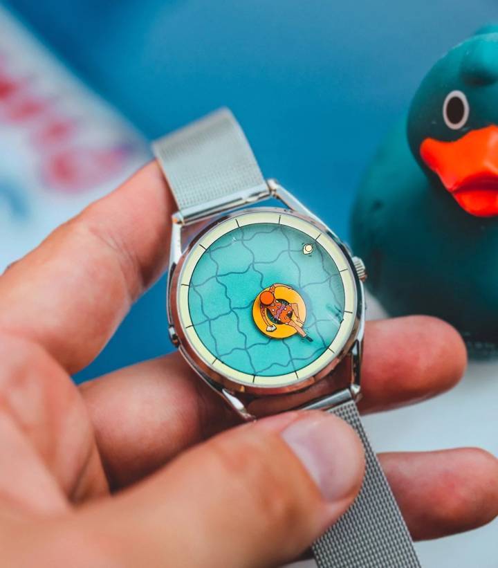 'Una tarde perfectamente inútil' no tiene números ni marcadores de minutos. El dial presenta una vista de pájaro de una persona descansando en una piscina. La pierna estirada de la figura relajada indica la hora, mientras que un pequeño pato de goma flota para mostrar los minutos. Las líneas de los mosaicos actúan como marcadores de las horas alrededor del borde de la piscina.